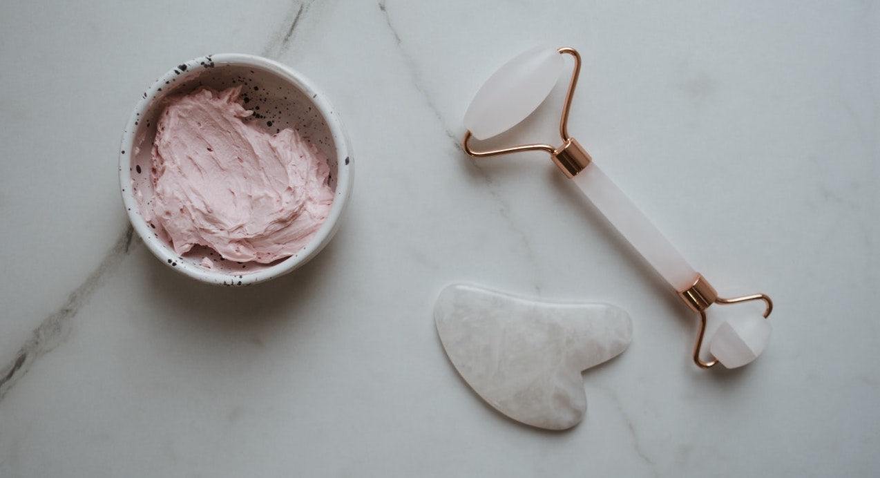 Bowl of pink face cream, rose quartz facial roller, and gua sha stone on marble surface.