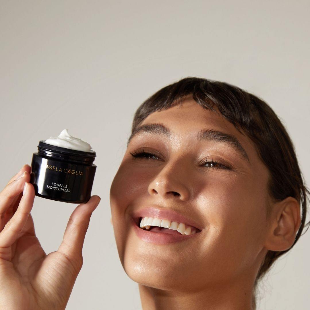 Smiling woman holding jar labeled 'ANGELA CAGLIA SOUFFLÉ MOISTURIZER' by her face