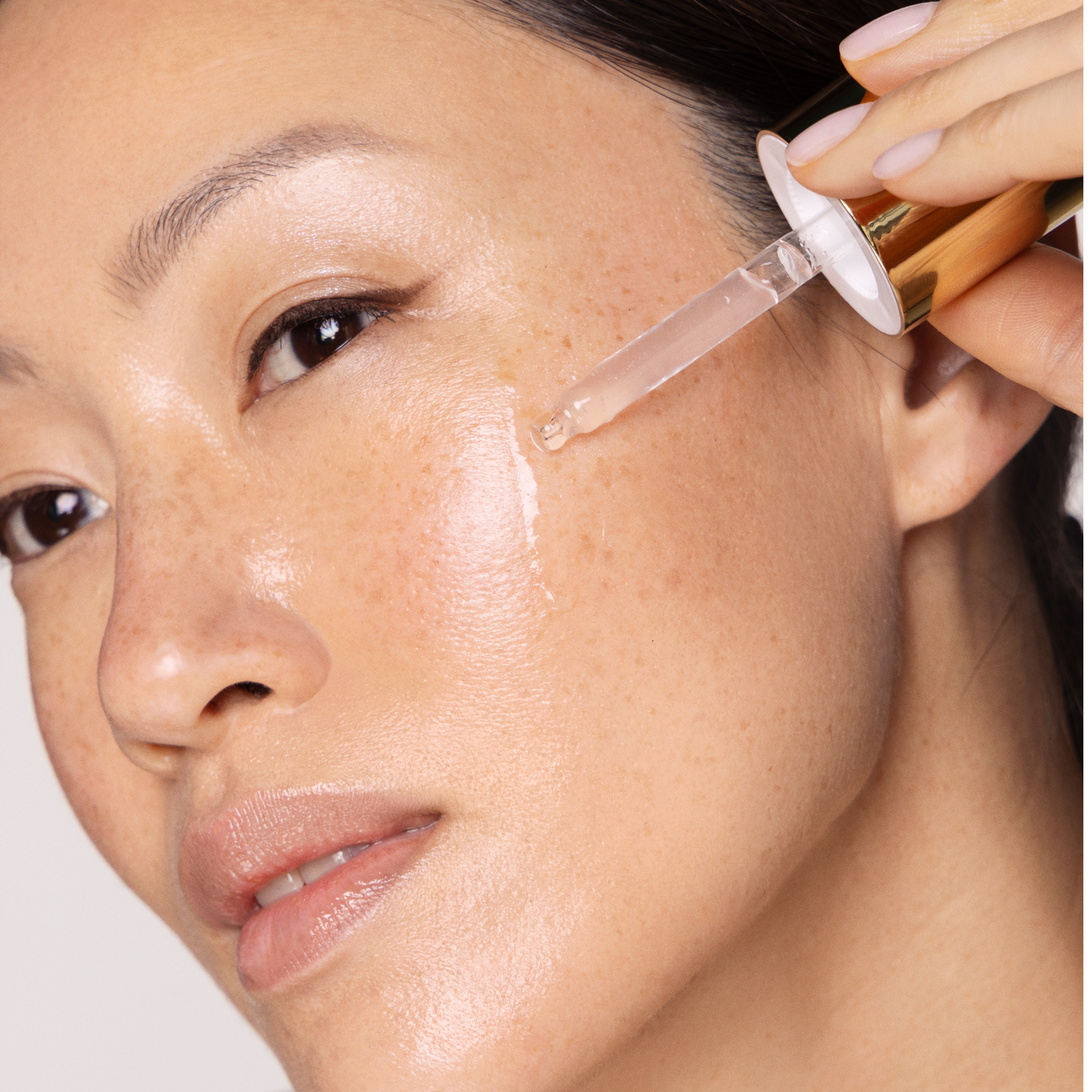 Person applying clear facial serum to cheek with a glass dropper, close-up on skin texture.