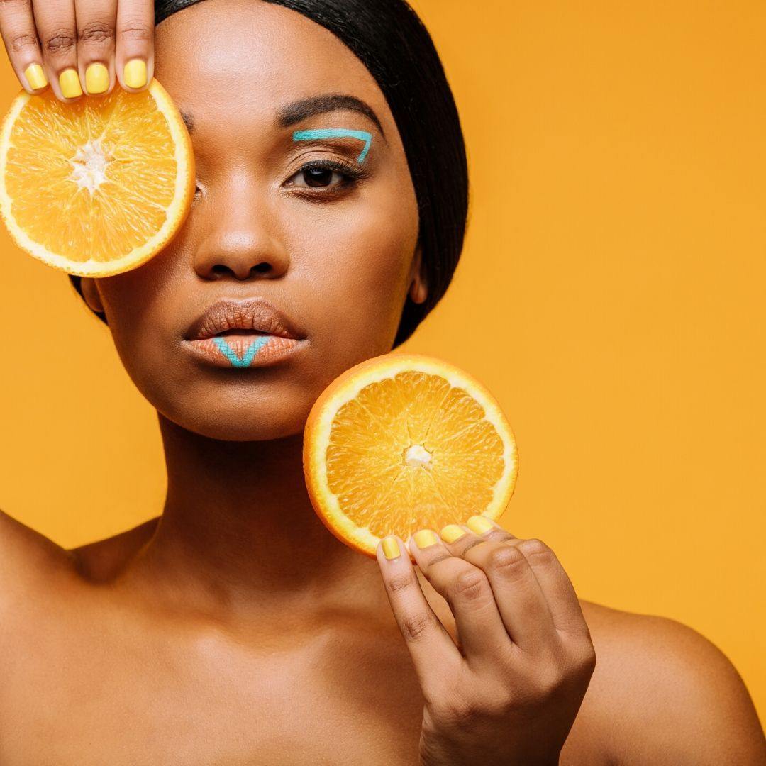 Woman with turquoise eye and lip makeup holding two orange slices, one covering her eye, on a yellow background.