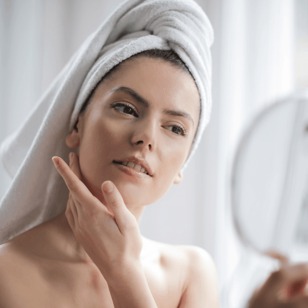 Young woman with towel-wrapped hair checking her face in a handheld mirror, touching her jawline.