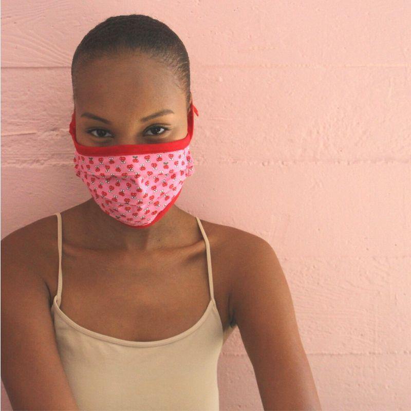 Woman with pulled-back hair wearing a pink cloth face mask with red floral print, beige camisole against a pink wall.