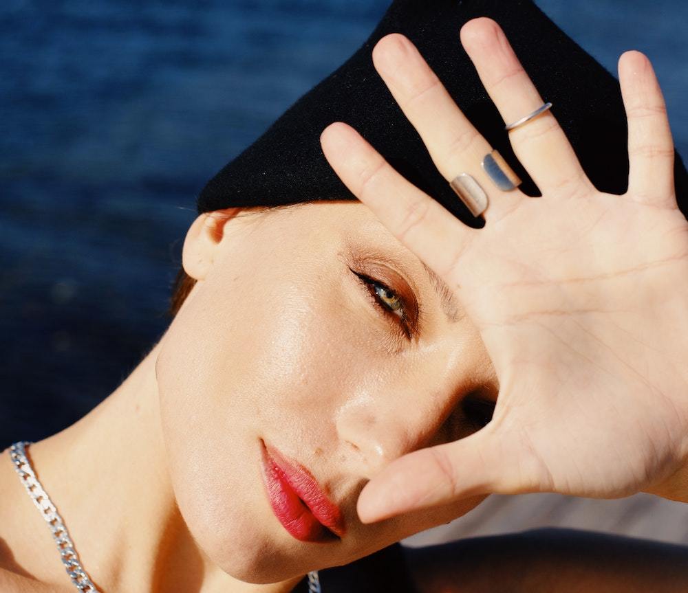 Woman wearing a black beanie shading her eyes with a hand, red lipstick and two rings visible