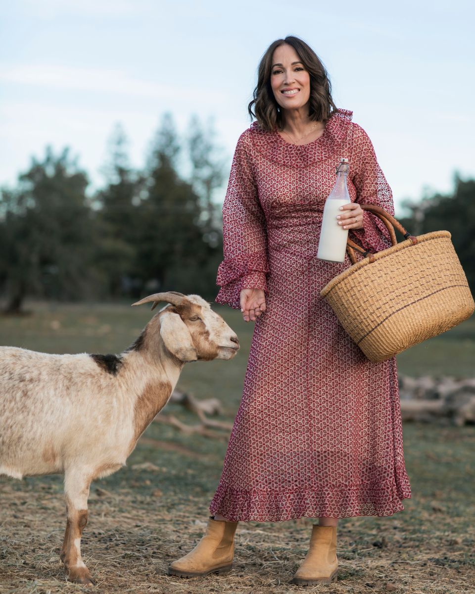 Smiling woman in patterned red dress holding a milk bottle and woven basket, standing beside a goat in a field