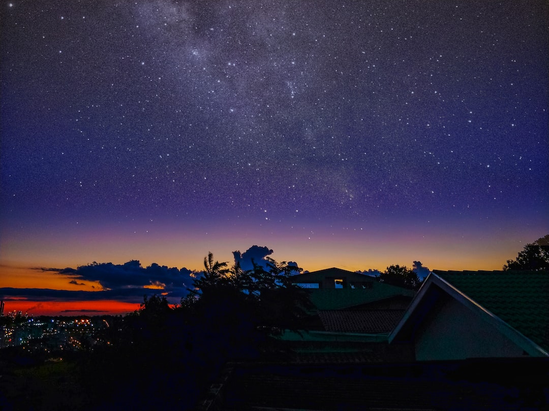 Star-filled night sky and Milky Way above rooftops at dusk, with orange-red sunset glow and distant city lights.