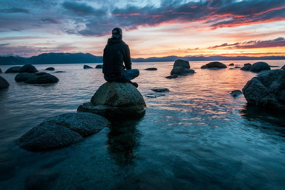 Person sitting on a rock at a lake, facing a colorful sunset over distant mountains