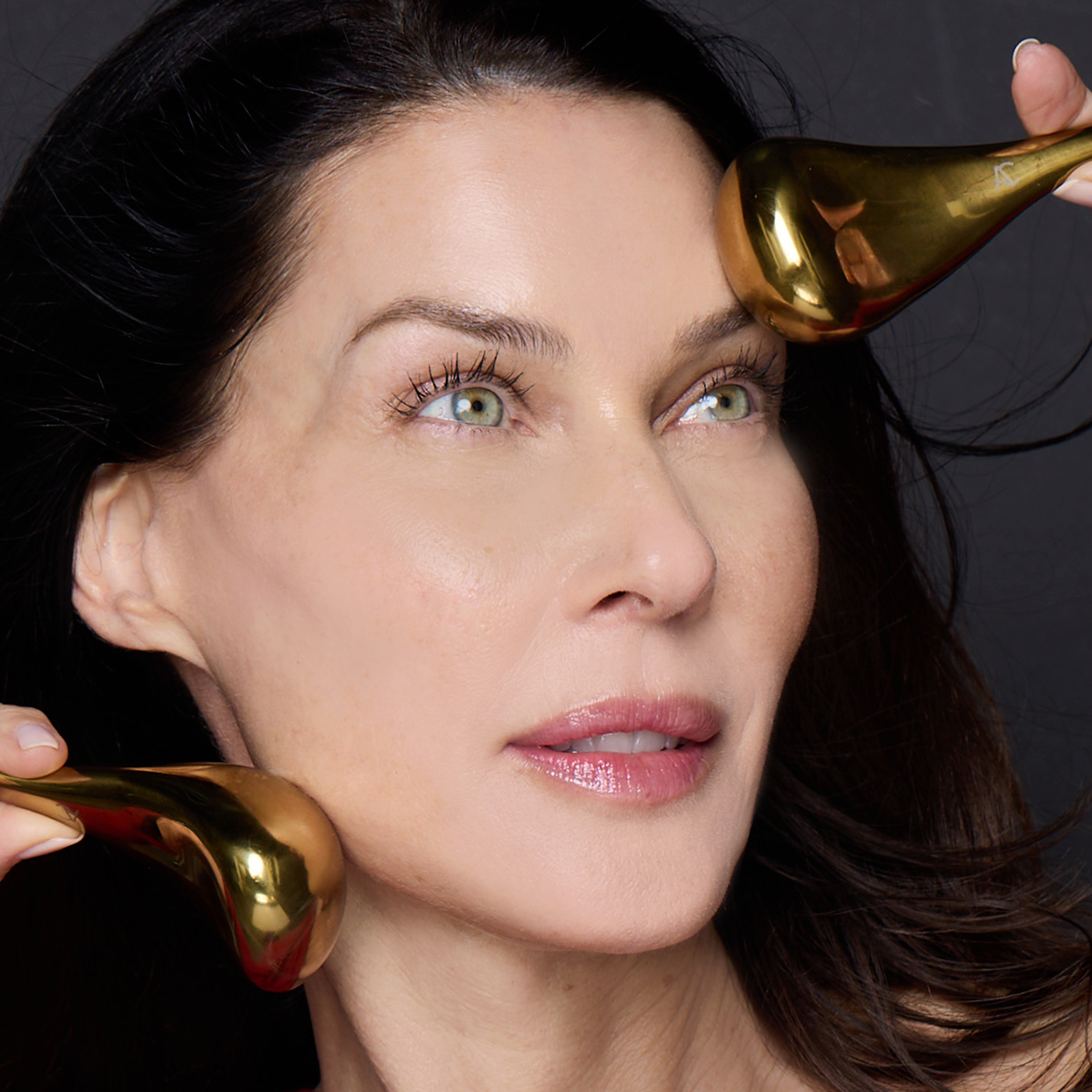 Brunette model holding gold-plated tool in each hand to side of face.