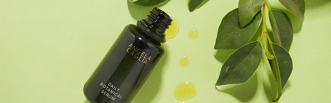 Dark green serum bottle labeled 'ANGELA CAGLIA DAILY BOTANICAL SERUM', oil drops and green leaves on pale green background
