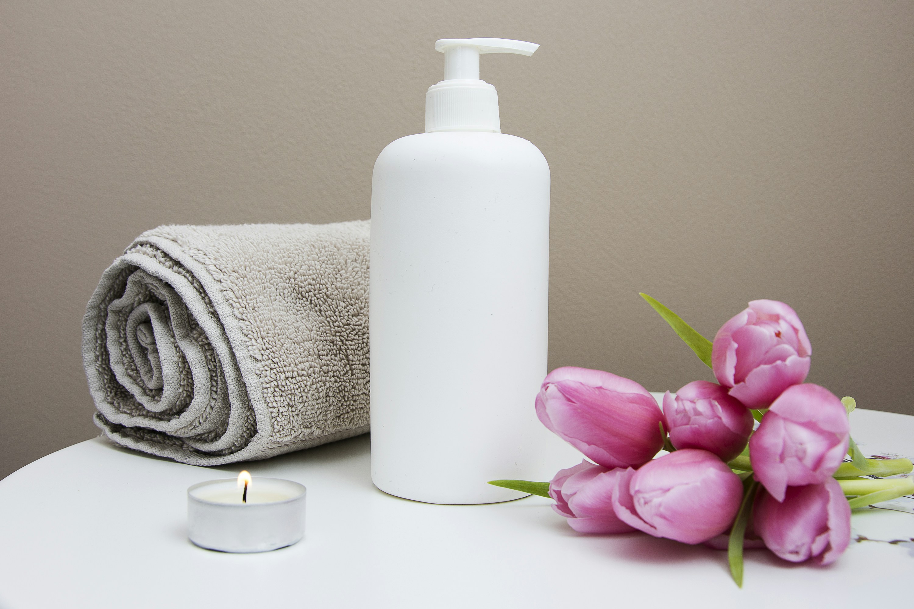 Spa scene with white pump bottle, rolled beige towel, lit tealight, and pink tulips on a white table